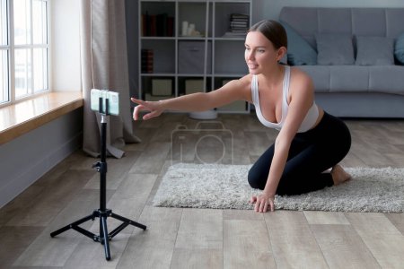 Foto de Mujer sentada en el suelo en pantalones de yoga grabando un video de fitness en línea con su teléfono y trípode, sonriendo a la cámara en una sala de estar - Imagen libre de derechos
