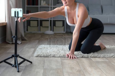 Téléchargez les photos : Femme assise sur le sol en pantalon de yoga enregistrant une vidéo de fitness en ligne avec son téléphone et son trépied, souriant à la caméra dans un cadre de salon - en image libre de droit