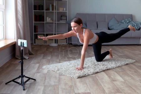 Foto de Mujer con leggins negros y un top blanco hace yoga en casa en la alfombra, grabando un video de fitness en línea para su audiencia con un smartphone de pie en un trípode - Imagen libre de derechos