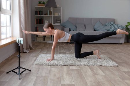 Photo for Woman in black leggings and a white top does yoga at home on the carpet, recording an online fitness video for her audience with a smartphone standing on a tripod - Royalty Free Image