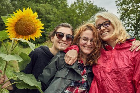 Photo for Girls posing for picture with sunflower smiling having fun, rainy overcast weather on summer vacation tour - Royalty Free Image