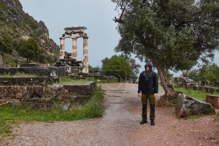Foto de Antiguas ruinas del templo en Delfos, Grecia, turista empapado en la lluvia, hombre no mirando demasiado entusiasta, solo, lejos del grupo, viajero solo - Imagen libre de derechos