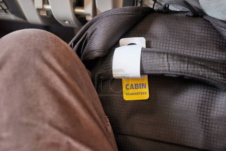 Téléchargez les photos : Sac cabine sur un vol avec étiquette pour bagage à main garantie ajustement entre les jambes et sous le siège à l'avant - en image libre de droit