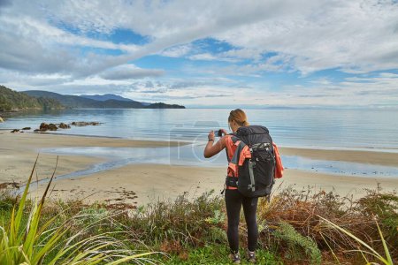 Photo for Woman hiking with backpack arriving to sandy beach in New Zealand, Stewart Island, North West Circuit. Taking a picture with cell phone camera - Royalty Free Image