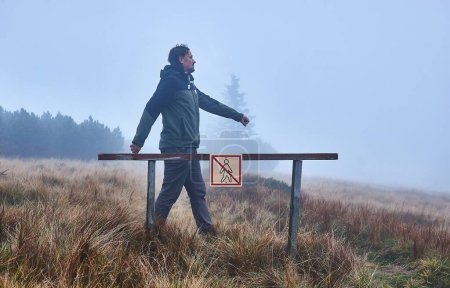 Man walking around barrier in a closed protected forest area, making fun of the figure on the no entry sign mimicking its shape, disobeying the rules. Cool foggy weather, twilight in the mountains.