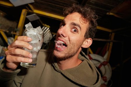 Kerl hält ein Glas voll Eiswürfel auf einer betrunkenen Garagenparty und beißt Eis zwischen die Zähne