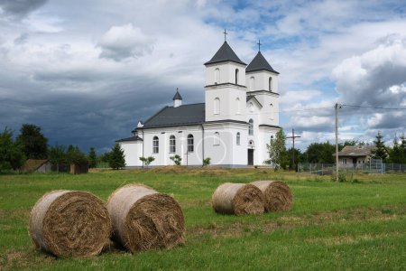Photo for View of the old ancient catholic Trinity Church and hay stacks on field, Yushkovichi village, Baranovichi district, Brest region, Belarus. - Royalty Free Image