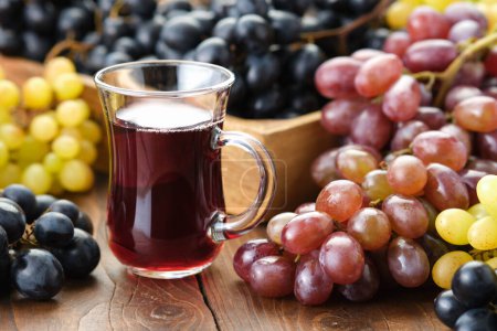 A glass cup of grape juice or wine. Black, green and purple grapes on table.