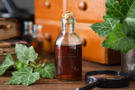 Photo for Bottle of ivy leaf syrup or cough tincture. Ivy leaves extract bottle. Wooden medicine cabinet with apothecary remedies, old books on background. Alternative herbal medicine. - Royalty Free Image