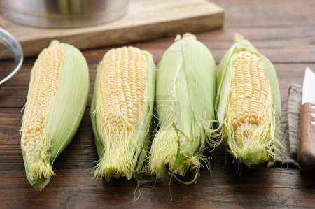 Foto de Mazorcas de maíz dulce crudo para cocinar y cuchillo de cocina en la mesa de la cocina, olla de cocina en el fondo. - Imagen libre de derechos