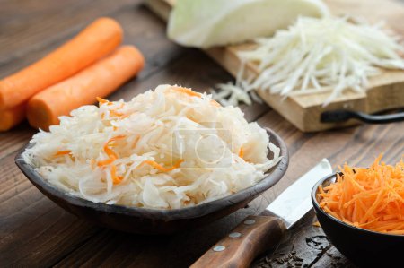 Photo for Bowl of sour cabbage, pickled sauerkraut. Fermented cabbage, coleslaw salad. Chopped cabbage on a cutting board and carrots for making sauerkraut on background. Healthy food, diet food. - Royalty Free Image