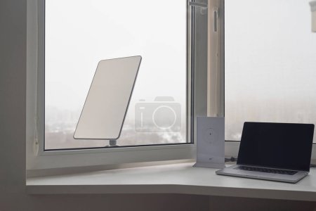 Photo for KYIV, UKRAINE - JANUARY 24, 2023: Starlink wi-fi router and laptop on windowsill at home with antenna behind window. - Royalty Free Image