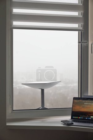 Photo for KYIV, UKRAINE - JANUARY 24, 2023: Starlink antenna receiving signal behind window, laptop with Starlink software and smartphone on windowsill. - Royalty Free Image