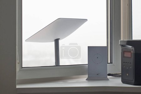 Photo for KYIV, UKRAINE - JANUARY 24, 2023: Close-up of Starlink antenna receiving signal behind window, wi-fi router and Ecoflow generator working on windowsill at home. - Royalty Free Image