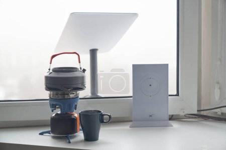 Photo for KYIV, UKRAINE - JANUARY 24, 2023: Close-up of Starlink antenna and wi-fi router on windowsill with portable burner stove with kettle and cup of tea beside. - Royalty Free Image