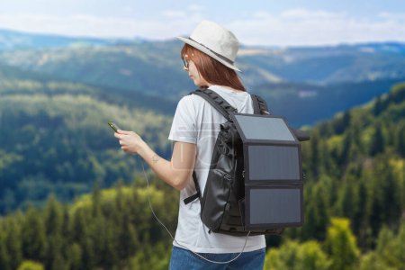 Foto de Mujer joven con teléfono móvil en manos conectadas a la mochila con batería solar portátil en el fondo del paisaje de las montañas. Concepto de viajes y energías renovables. - Imagen libre de derechos