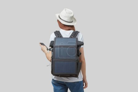 Photo for Girl carrying backpack with flexible solar panel attached to smartphone in her hands, isolated on light gray background. Travel and alternative energy concept. - Royalty Free Image
