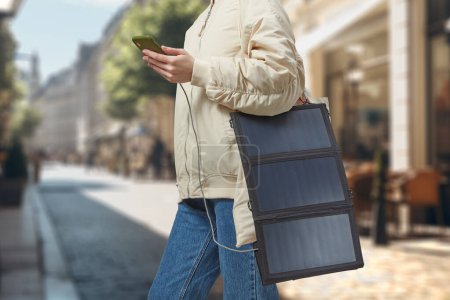 Téléchargez les photos : Femme méconnaissable avec batterie solaire flexible tenant smartphone en arrière-plan d'une ville. Concept de voyage et d'énergie alternative. - en image libre de droit