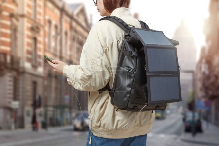 Téléchargez les photos : Fille avec téléphone portable dans les mains et batterie solaire portable sur le sac à dos voyageant dans une ville. Concept de voyage durable. - en image libre de droit