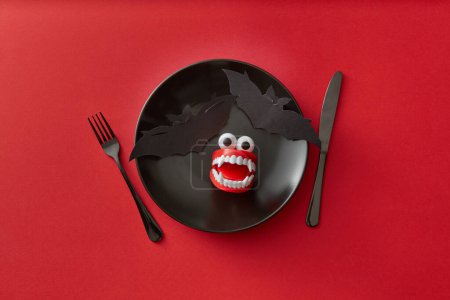 Photo for A black plate with a bat and vampire teeth on it, on a red background. A knife and fork on each side add to the horror theme. A perfect plate setting for a Halloween meal. - Royalty Free Image