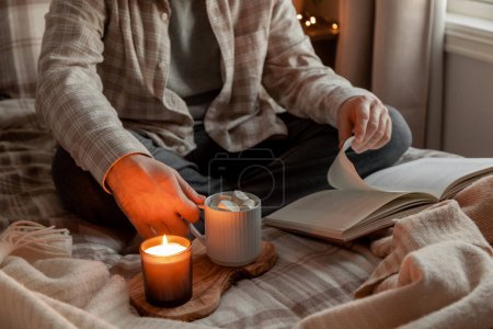 ein kaukasischer Mann entspannt sich zu Hause, zündet Kerzen an, trinkt Kaffee und liest Buch im Bett