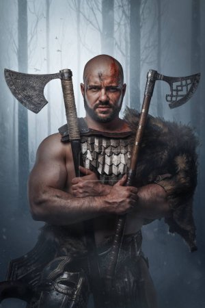 Portrait of ancient viking with fur and horned helmet holding axes in cross in winter woods.