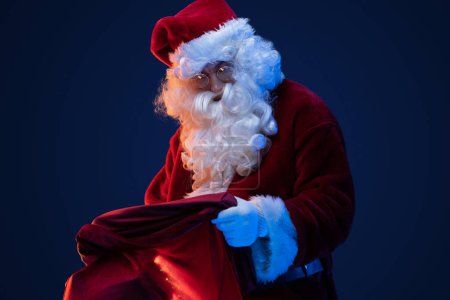 Foto de Retrato de Papá Noel viejo con gafas y bolsa con regalos sobre fondo oscuro. - Imagen libre de derechos