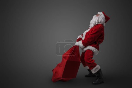 Photo for Studio shot of funny santa claus dragging big bag with gifts against gray background. - Royalty Free Image