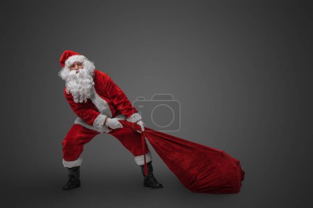 Photo for Shot of bearded santa with big bag stuffed with gifts against grey background. - Royalty Free Image