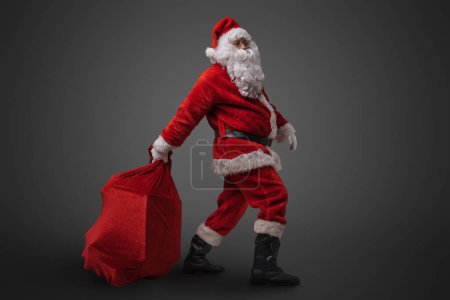 Photo for Studio shot of funny santa claus dragging big bag with gifts against gray background. - Royalty Free Image