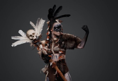 Shot of painted dark witch with fur and staff against grey background.
