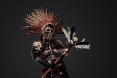 Foto de Foto de bruja azteca del pasado con tocado de plumas y máscara que sostiene al personal con cabeza de cráneo. - Imagen libre de derechos