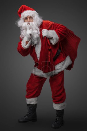 Photo for Photo of santa claus with bag and his finger at his lips against gray background. - Royalty Free Image