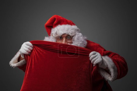 Photo for Shot of funny santa with glasses and hat hiding behind bag against grey background. - Royalty Free Image