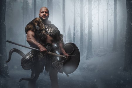 Photo for Studio shot of handsome ancient viking holding axe and shield in forest in snowfall. - Royalty Free Image