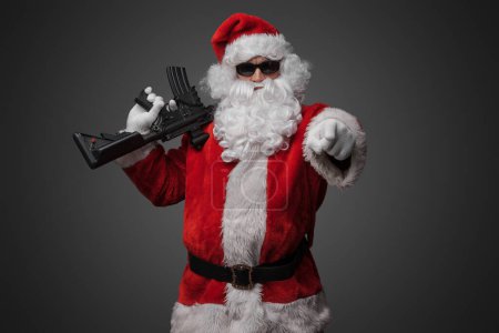 Téléchargez les photos : Prise de vue du père Noël cool tenant fusil sur son épaule pointant son doigt vers la caméra. - en image libre de droit