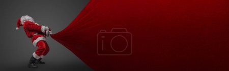 Photo for Shot of xmas santa dressed in red costume pulling huge bag with christmas presents. - Royalty Free Image