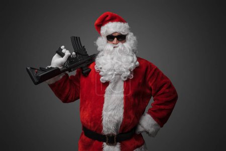 Photo for Shot of christmas santa claus with red costume and rifle against gray background. - Royalty Free Image