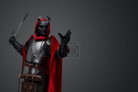 Foto de Portrait of member of dark cult dressed in black mask and red robe holding sword. - Imagen libre de derechos