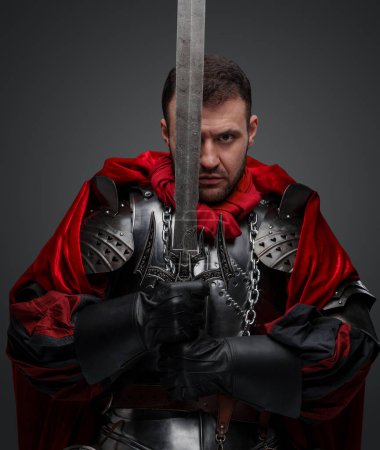 Foto de Tiro de caballero oscuro vestido con armadura de acero que sostiene la espada contra el fondo gris. - Imagen libre de derechos