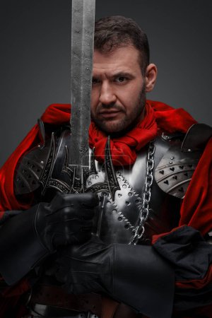 Foto de Tiro de caballero oscuro vestido con armadura de acero que sostiene la espada contra el fondo gris. - Imagen libre de derechos