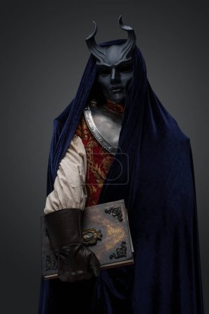 Foto de Tiro de cultista con máscara de cuernos y capa azul oscuro sobre fondo gris. - Imagen libre de derechos