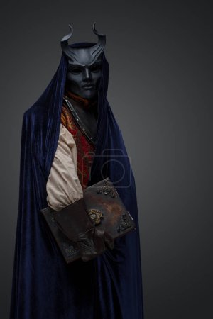 Foto de Tiro de cultista oscuro vestido con máscara de cuernos y bata sosteniendo libro. - Imagen libre de derechos