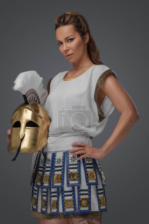 Foto de Estudio de disparo de guerrera griega vestida con armadura ligera y sosteniendo casco dorado. - Imagen libre de derechos