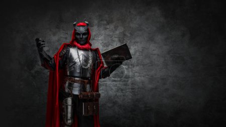 Foto de Shot of mysterious cultist dressed in steel armor and red mantle holding book. - Imagen libre de derechos