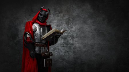 Téléchargez les photos : Shot of mysterious cultist dressed in steel armor and red mantle holding book. - en image libre de droit