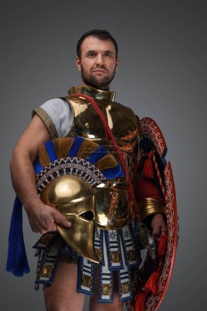 Foto de Studio shot of handsome greek warlord holding golden helmet against grey background. - Imagen libre de derechos