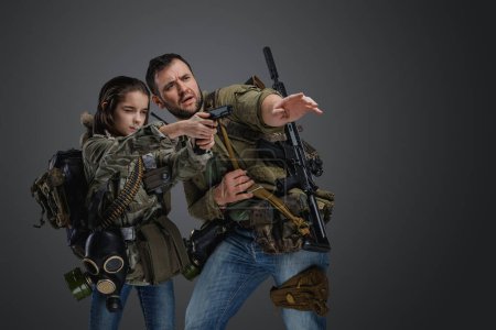 Téléchargez les photos : Portrait d'un homme entraînant une petite fille à tirer sur une arme de poing dans le cadre d'une post-apocalypse. - en image libre de droit