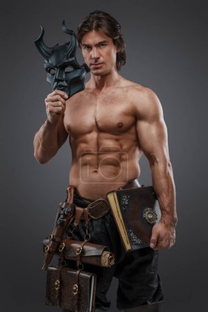 Shot of topless cultist man with muscular body holding mask against gray background.