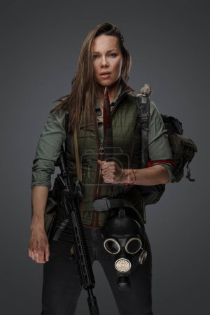 Banditenfrau mit blutigem Messer in postapokalyptischer Umgebung. Hochwertiges Fotoporträt einer Mörderin mit Pistole und Messer, die einen Rucksack vor grauem Hintergrund trägt.